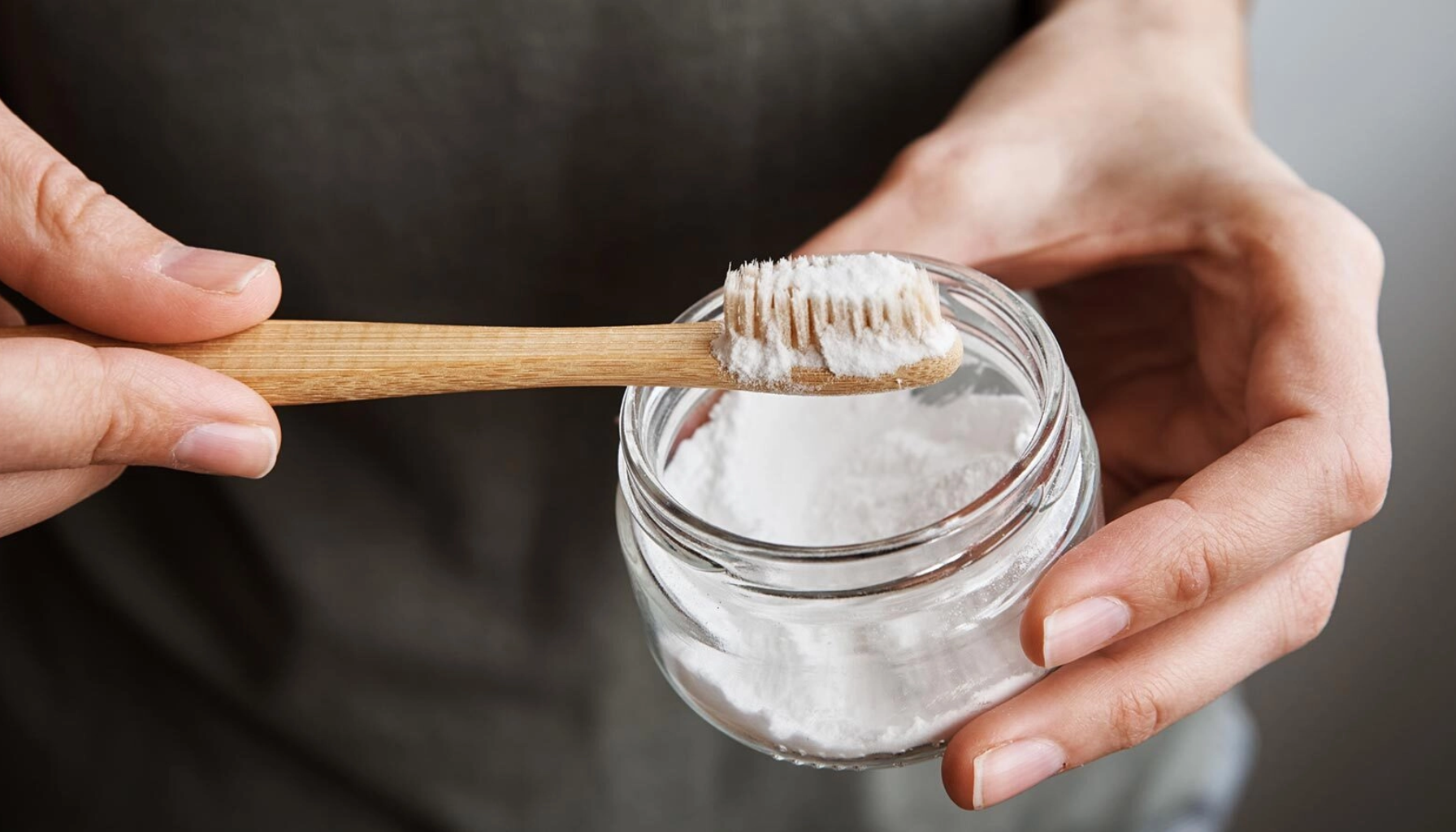 Person holder glass med natron og bambustannbørste, viser naturlig metode for tannbleking og munnhygiene.