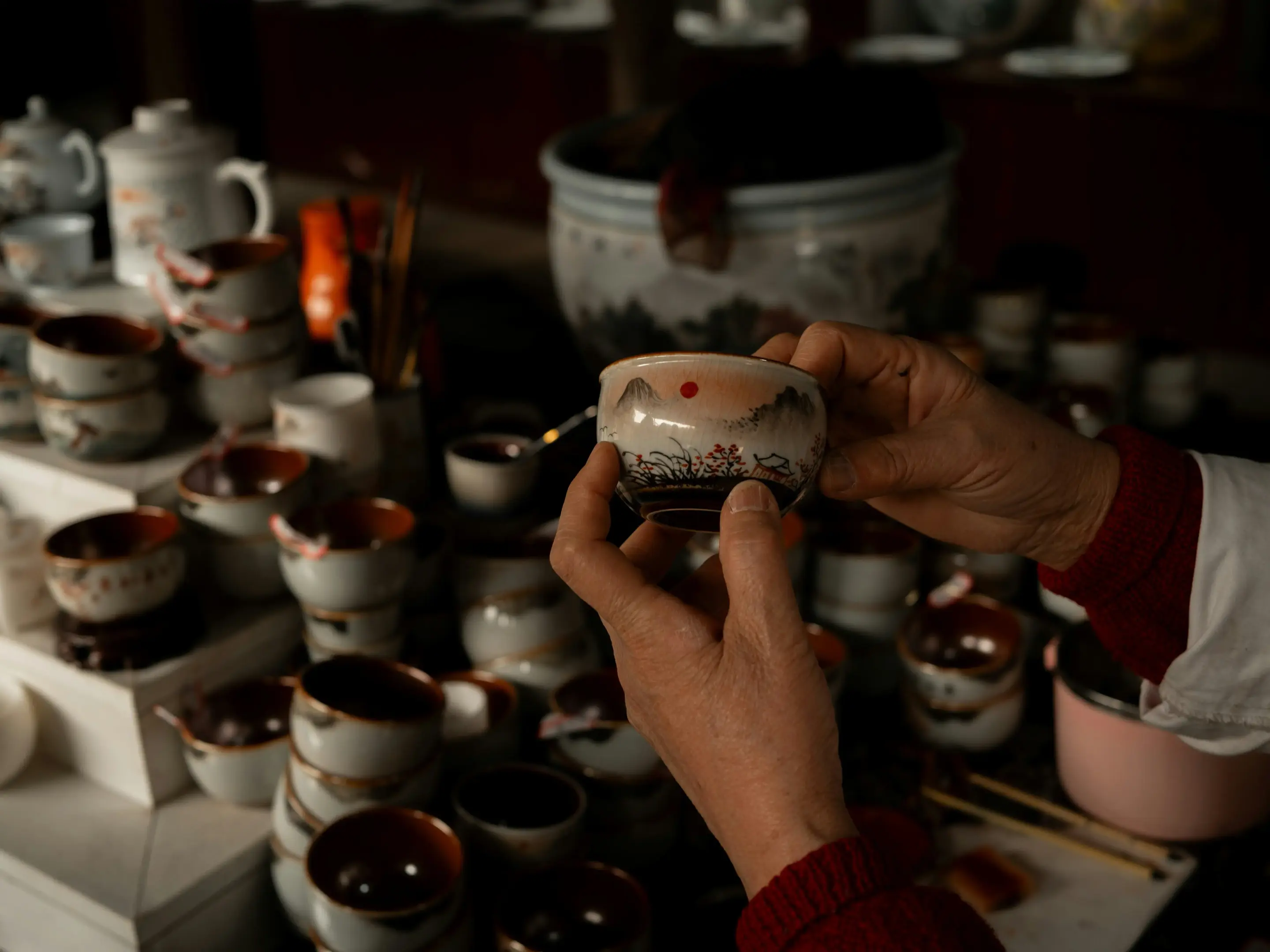 A pair of hands holds up a small, ornate ceramic tea cup featuring a painted landscape with a prominent red sun. The background is filled with stacks of similar traditional pottery and teaware in a dimly lit workshop or shop setting.