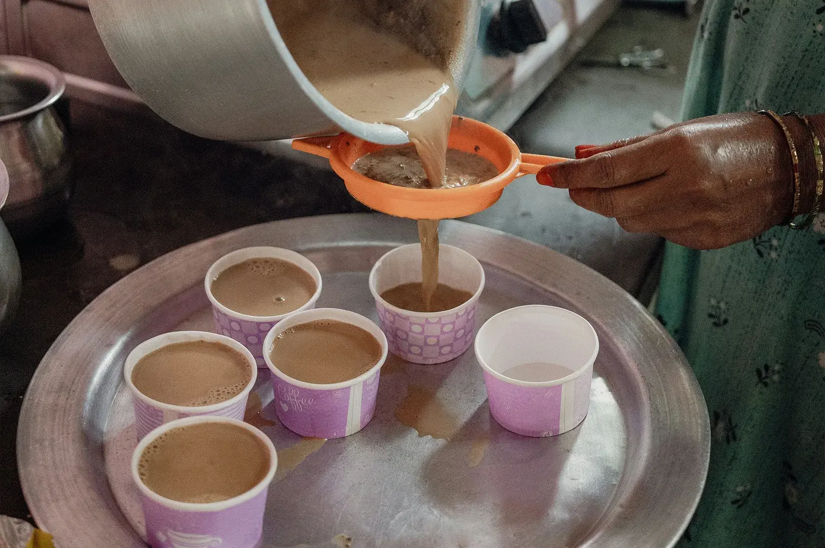A person’s hand is seen straining hot, milky masala chai through a small orange plastic sieve from a large metal pot into several paper cups arranged on a metallic tray, illustrating the preparation process in a busy kitchen setting.