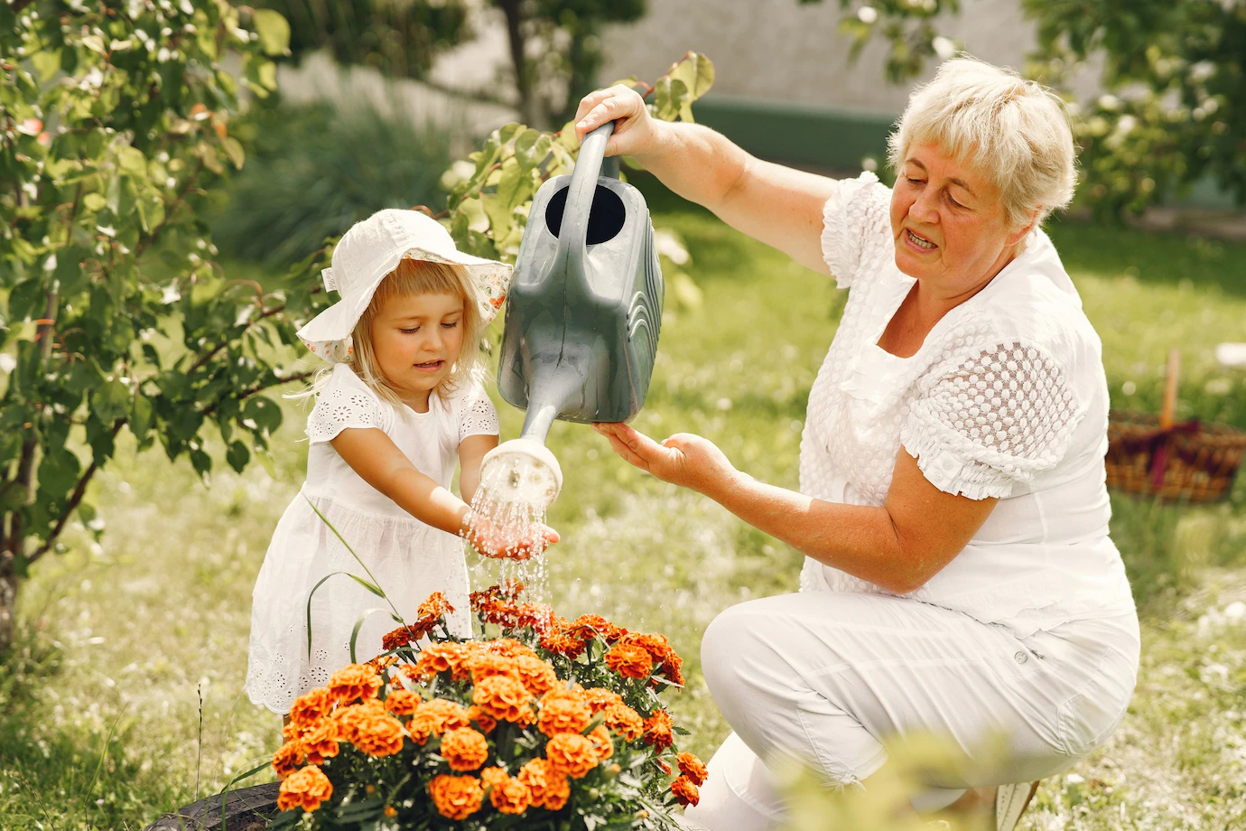 An elderly lady, in a garden, is watering some flowers with a watering can. A young girl is nearby and is holding her hands out under the water