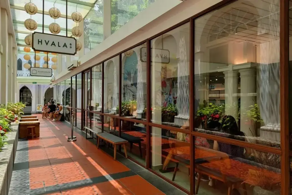 In the image, a bright, glass-walled corridor features the storefront of a cafe named Hvala, decorated with hanging globe lanterns. Patrons sit at tables behind large windows and on outdoor benches along a terracotta-tiled walkway bathed in natural light.