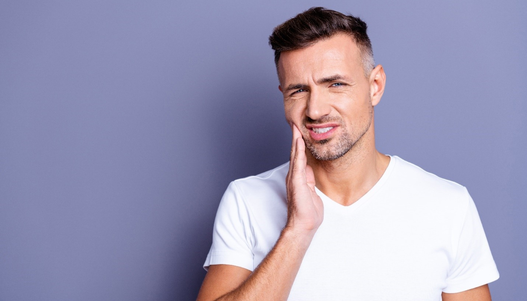 Mann i hvit t-skjorte holder seg på kinnet og uttrykker smerte, tydelig tegn på tannverk.