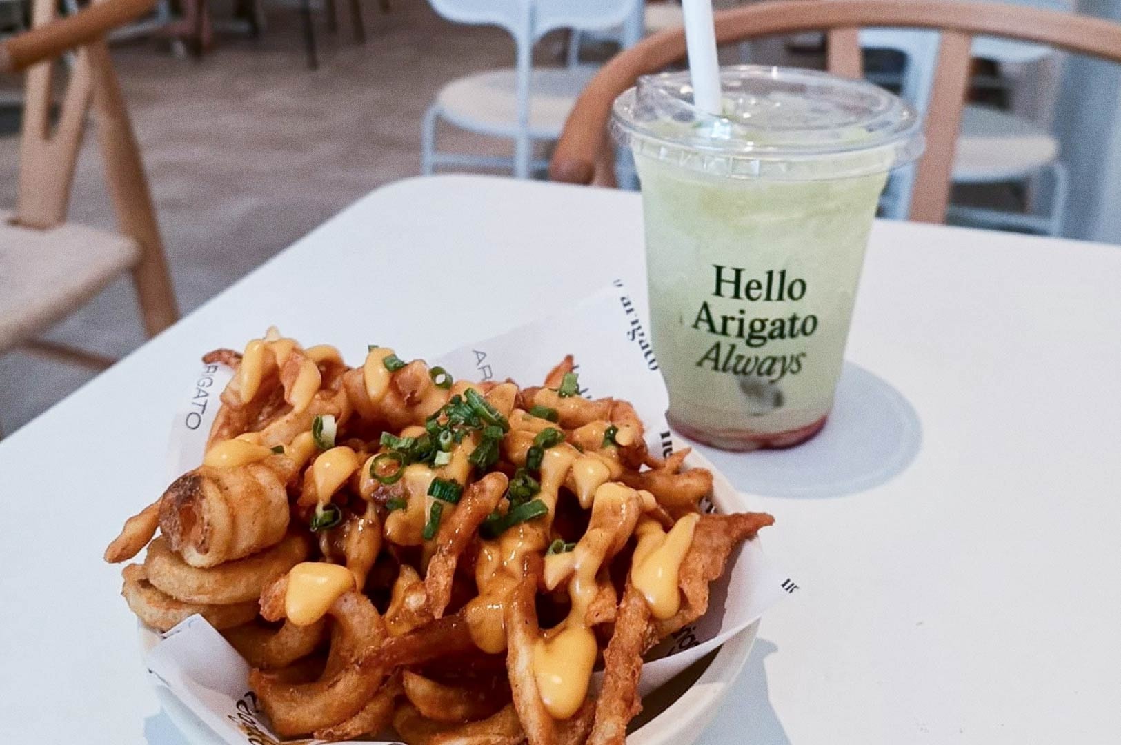 Bowl of cheesy curly fries topped with chopped green onions on branded paper reading ARIGATO, beside a clear plastic cup of layered green and red drink labeled Hello Arigato Always, set on a white table in a casual café.