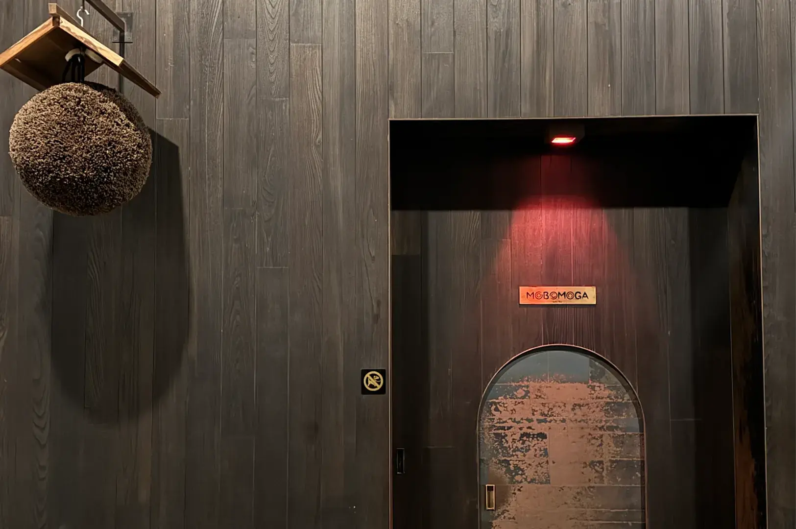 Entrance of Mobomoga sake bar featuring dark wooden paneled walls, a rounded doorway with red spotlighting, and a hanging cedar ball.
