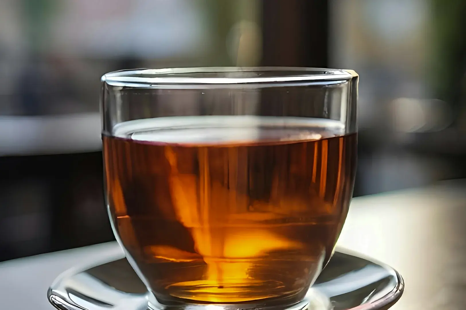 A straightforward, close-up profile shot features a clear, heavy-bottomed glass teacup filled with a rich, reddish-amber tea, resting on a matching clear glass saucer. The cup is positioned centrally against a completely blurred, out-of-focus background that hints at an outdoor or window-side setting with muted greens and greys, highlighting the clean lines of the glassware and the clarity of the steeped liquid.