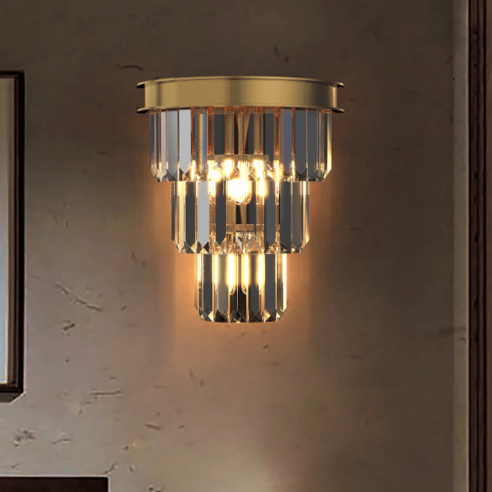 Elegant wall sconce featuring a tiered design of vertical crystal prisms mounted on a gold base, softly glowing with warm light against a textured wall.