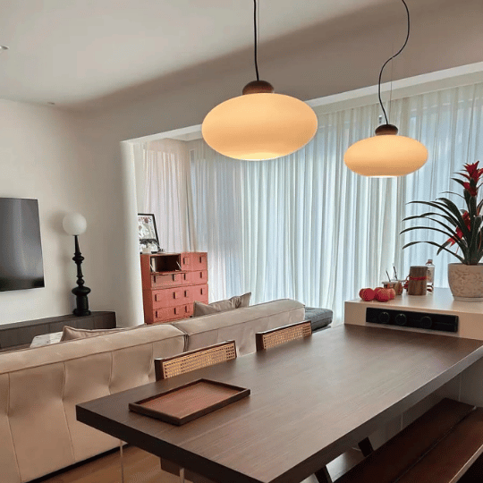 Modern living and dining area with a wooden table, beige sofa, and two warm-glow pendant lights hanging above, set against sheer white curtains.