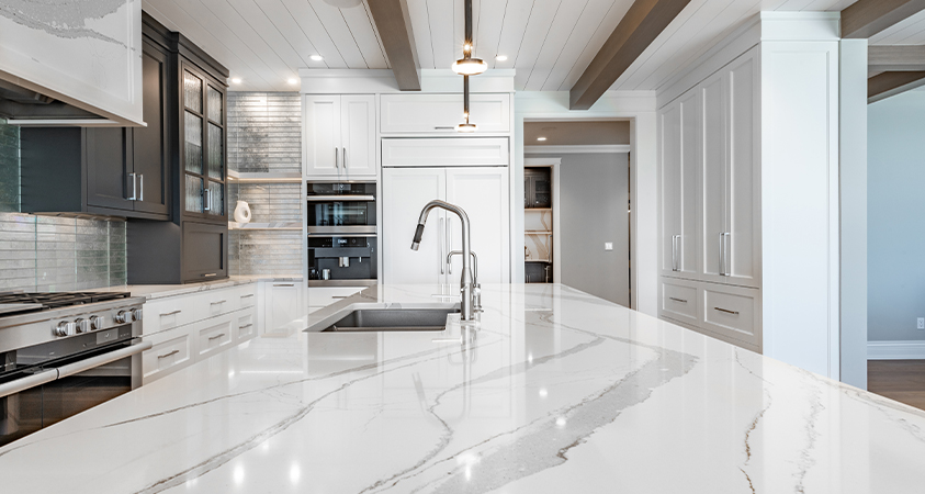 A smooth marble kitchen bench sparkles underneath some white down lights. The shiny white surface of the room is broken by dark wood cupboards and wood beams on the ceiling.