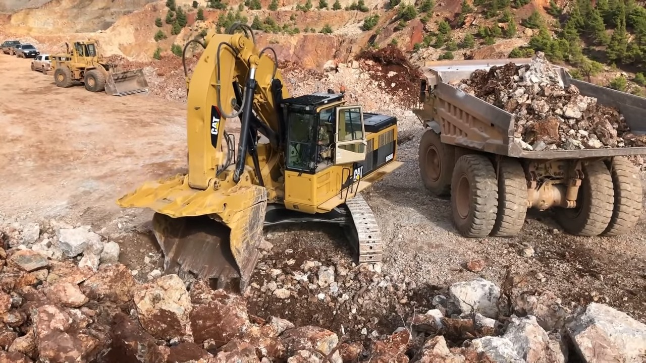 A yellow excavator with the CAT logo on its side, working on a construction site, digging through a pile of rocks and dirt. A large dump truck loaded with debris is parked nearby