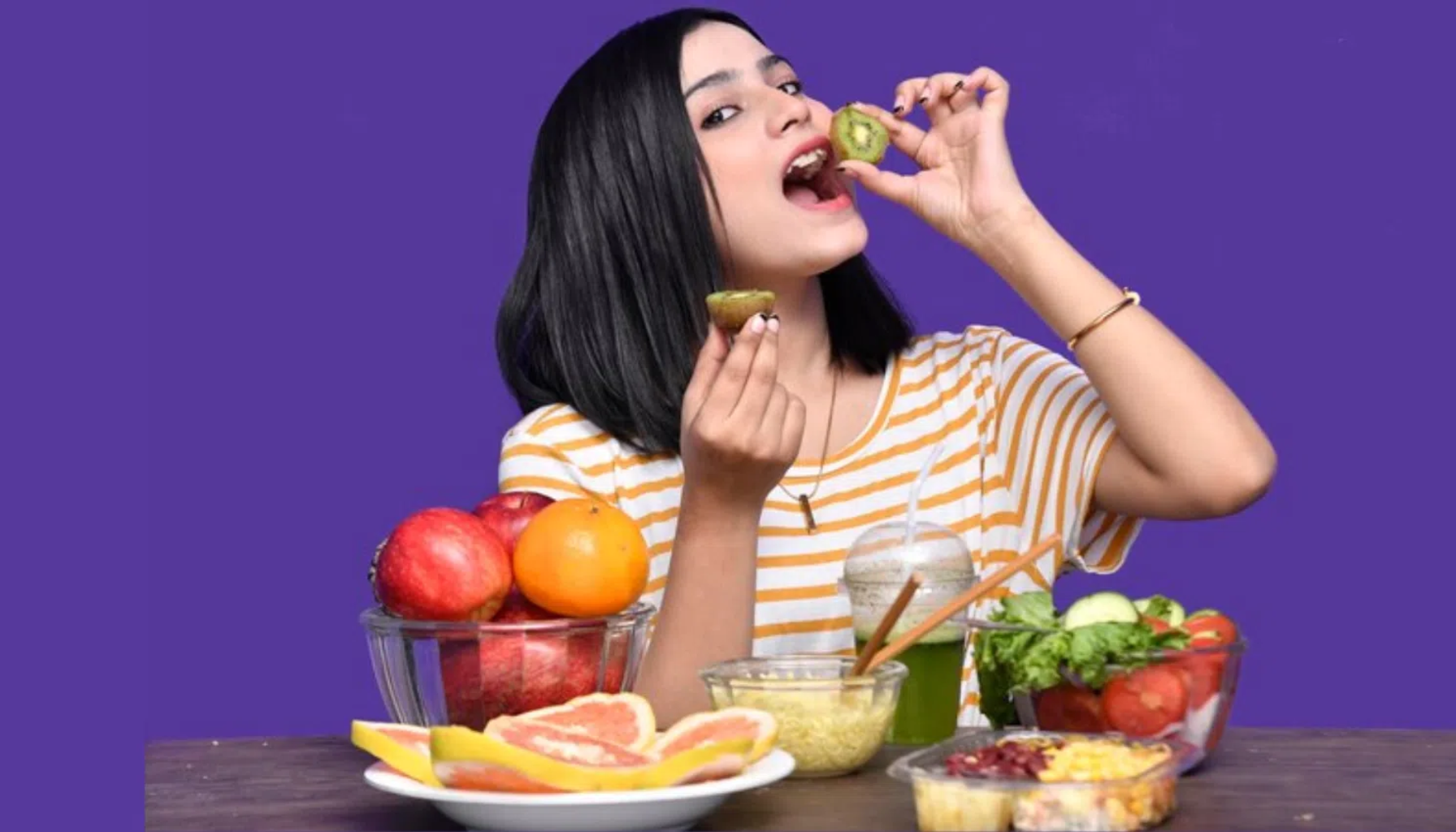 "Kvinne i stripete t-skjorte spiser kiwi foran et bord fullt av frukt og salat med lilla bakgrunn."
