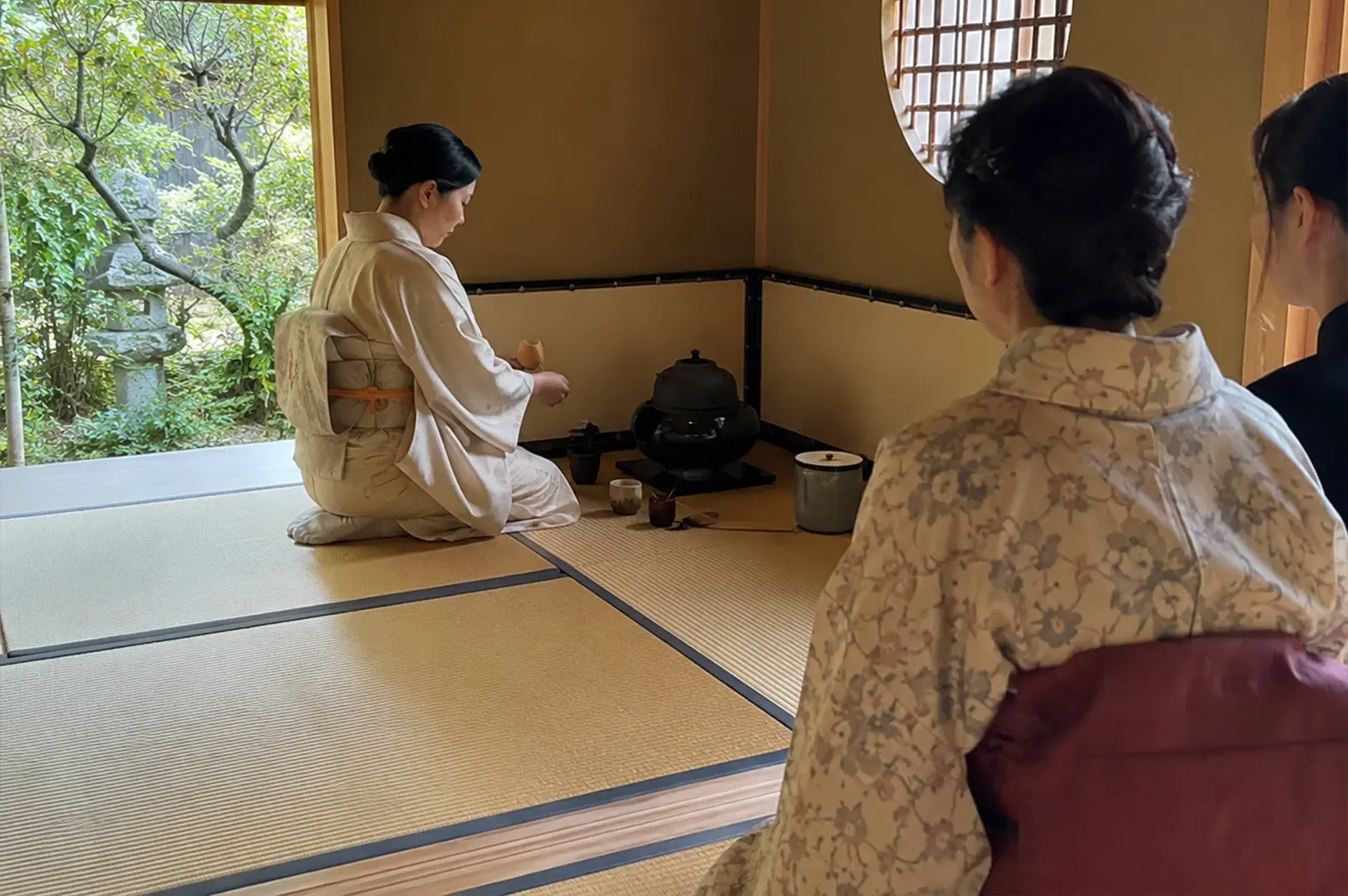 A host dressed in a cream-colored kimono is shown kneeling and preparing tea, while guests sit respectfully on the tatami mats to her right. The scene takes place in a minimalist room with paper-screened walls and a circular window, offering a view of a lush garden with a stone lantern outside. The composition highlights the communal yet meditative nature of the tea ceremony, framed within the traditional architecture of the space.