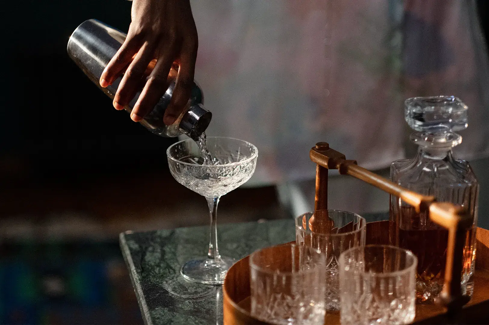 Person pouring a clear cocktail from a metal shaker into an ornate crystal coupe glass, with a bar tray holding whiskey glasses and a decanter nearby.