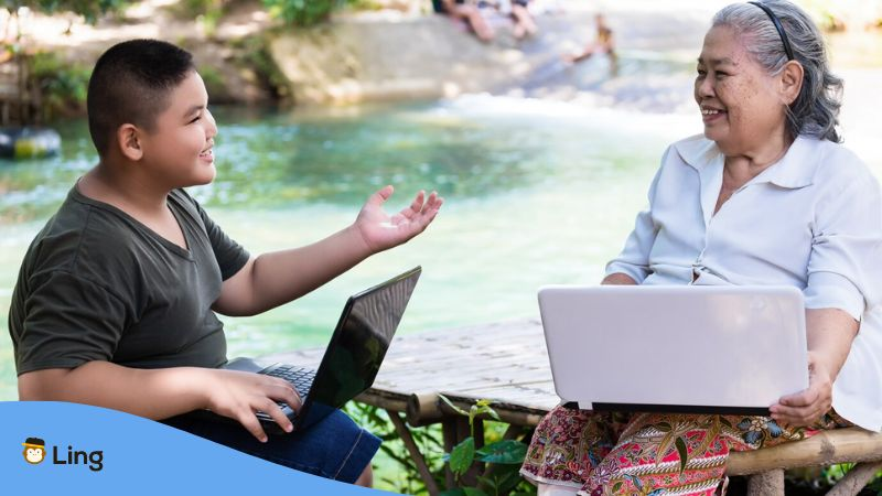 A photo of a boy talking to his grandmother using honorific nouns and Thai verbs outdoors with a river flowing behind them.