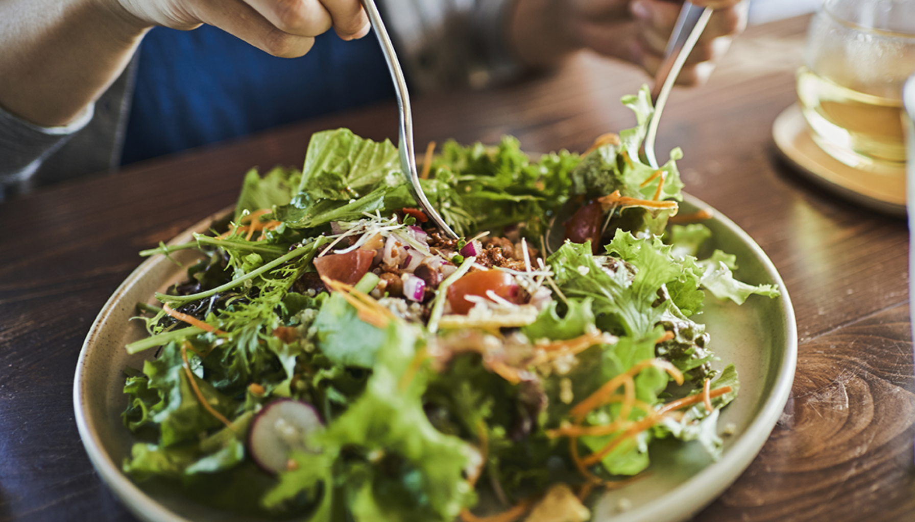 Person som spiser frisk grønn salat med tomater, urter og grønnsaker, løftet med to gafler over tallerkenen.