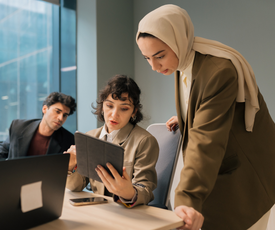 two women and one man gather around a desk to review something on a tablet and laptop