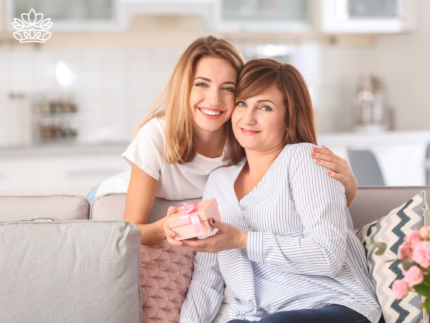 A young woman presents a gift to her mother, sharing a joyful moment on the sofa, reflecting the quality and thoughtfulness of the Johannesburg Gift Delivery Collection. Perfect for family celebrations such as birthdays and anniversaries, offering a variety of gifts and hampers. Fabulous Flowers and Gifts.