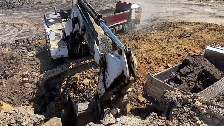 A large white excavator digging into the ground with its bucket, surrounded by piles of dirt and rocks, with a red and white dump truck in the background on a construction site