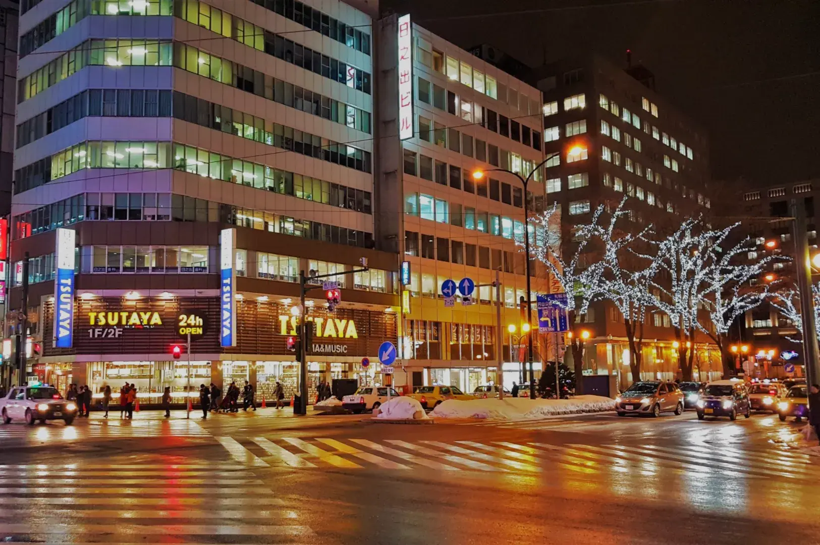 A street scene at night featuring illuminated buildings, including a Tsutaya store, with traffic and decorative lights on trees.