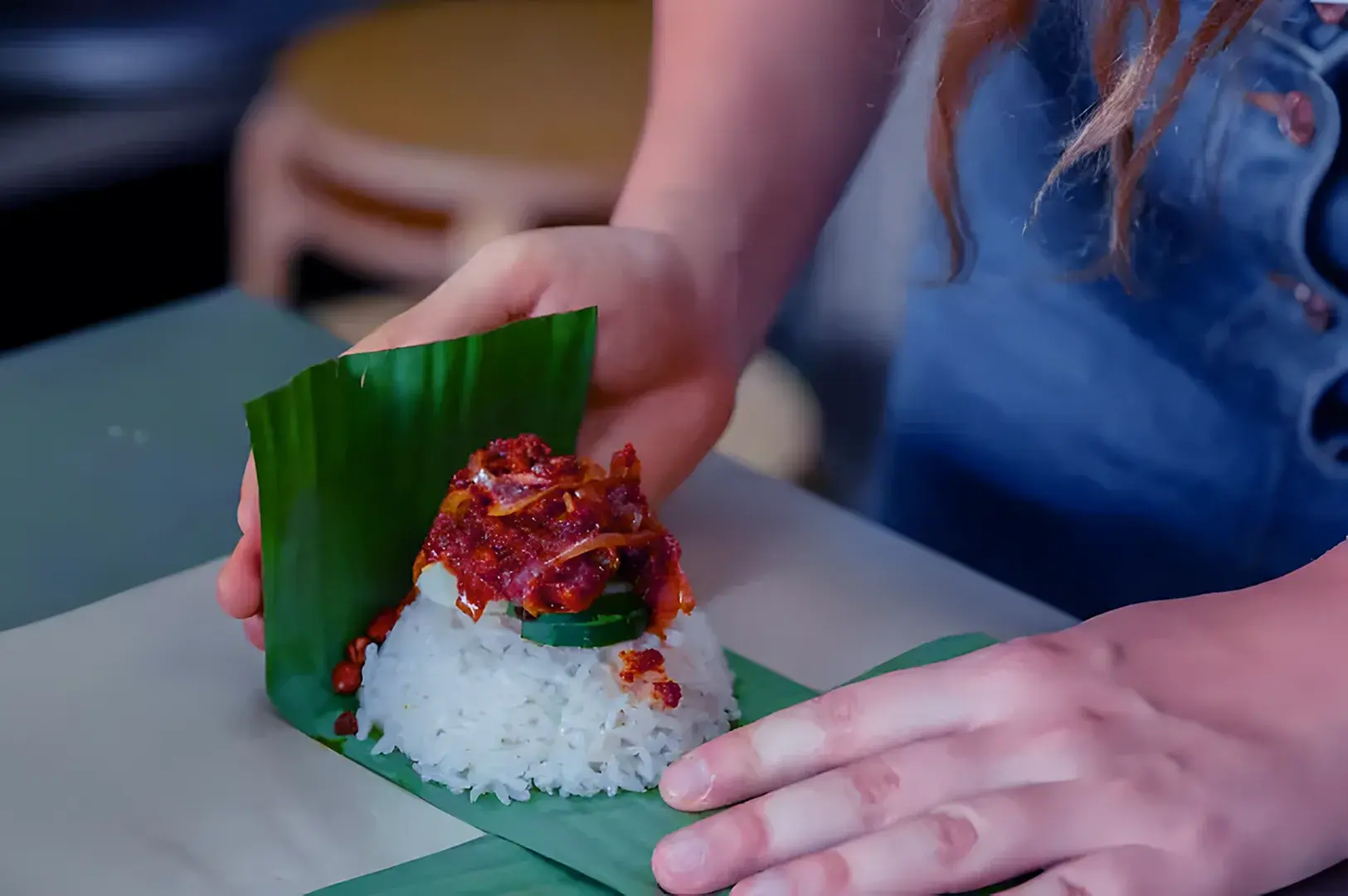 A person wraps a serving of rice and spicy sambal in a banana leaf. The scene conveys an authentic culinary experience, highlighting traditional food preparation.