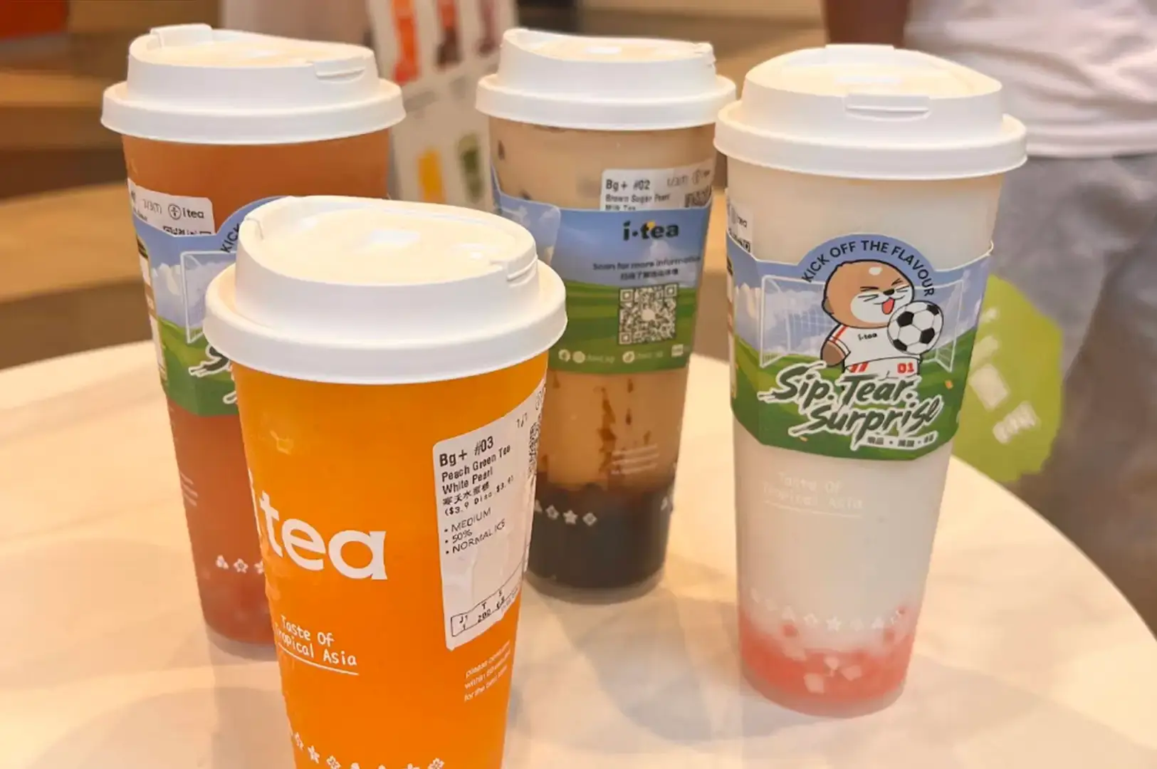 Four iTea beverages with white dome lids arranged on a white table, each wearing a green cardboard cup sleeve featuring a cartoon soccer-themed otter character. The drinks include an iced milk tea with brown sugar syrup streaks at the bottom, an orange fruit tea with white pearls, a reddish iced tea, and a creamy white coconut or milk drink layered with pink jelly pieces at the base.