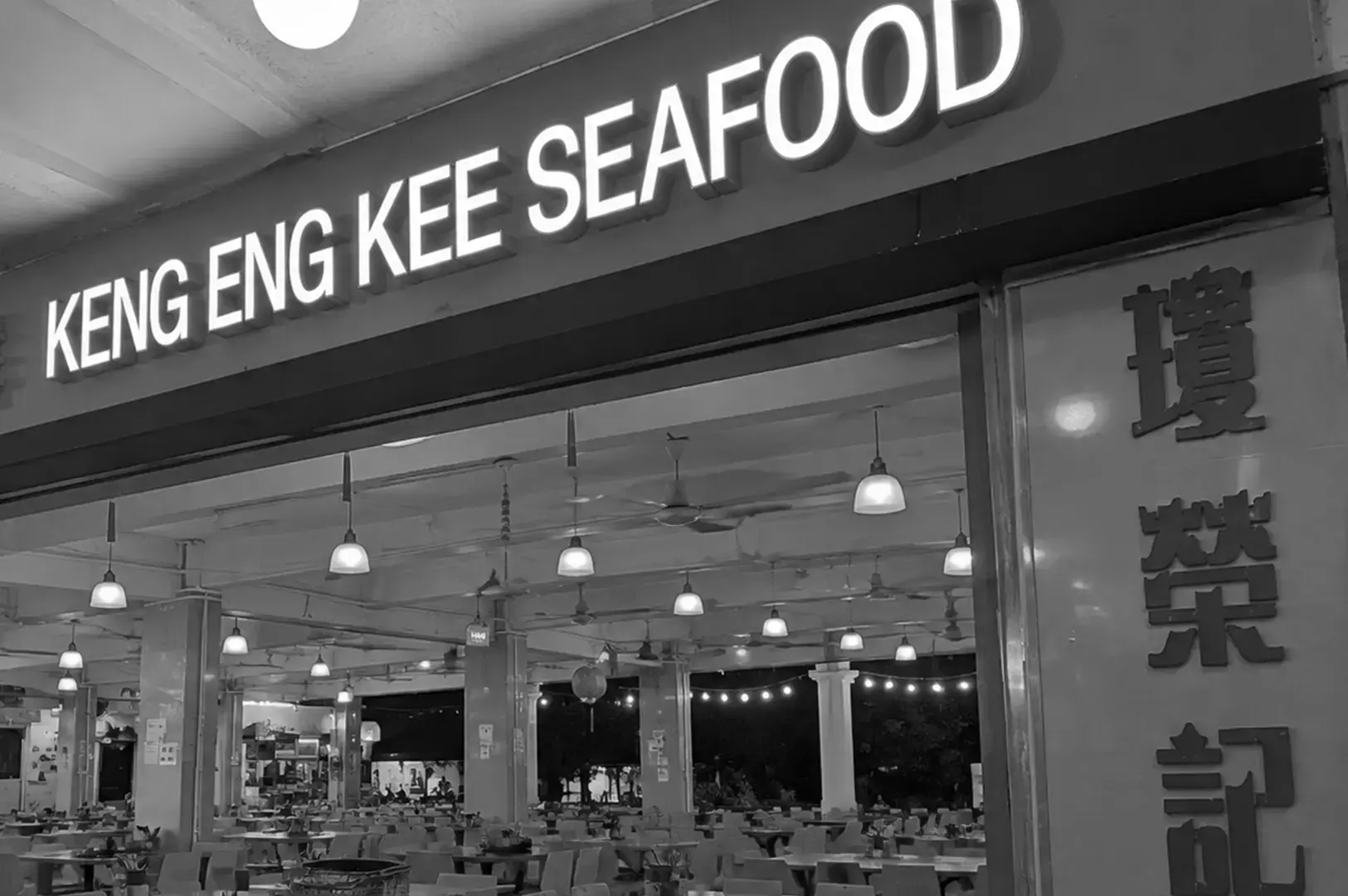 Keng Eng Kee Seafood restaurant entrance with its large illuminated sign above an open dining area filled with tables, chairs, hanging lights, and ceiling fans.