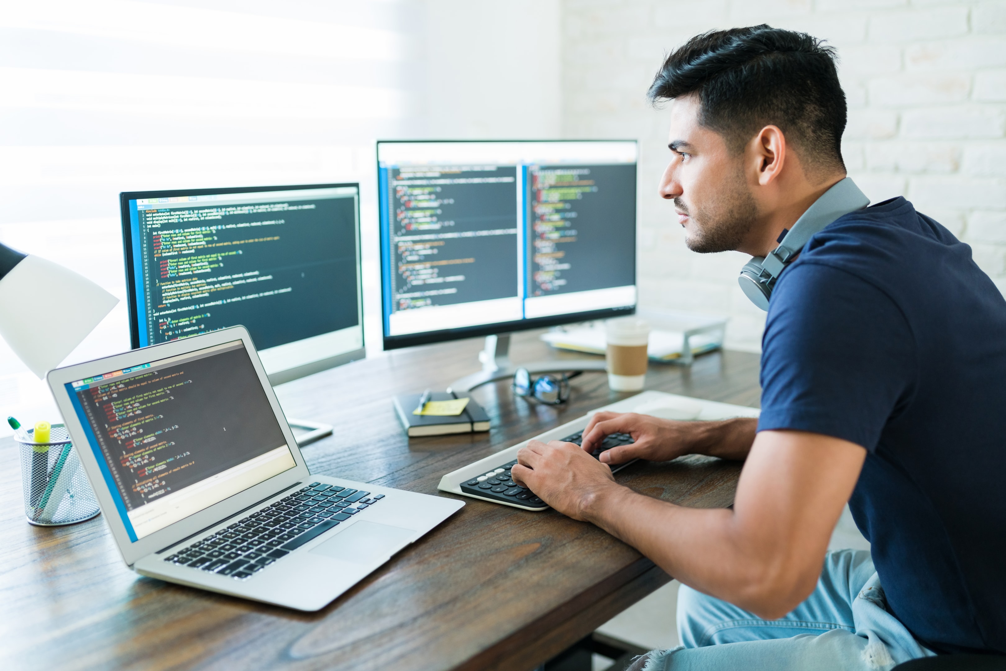 Focused male software developer working at a desk with a laptop and dual monitors displaying code, wearing headphones around his neck.