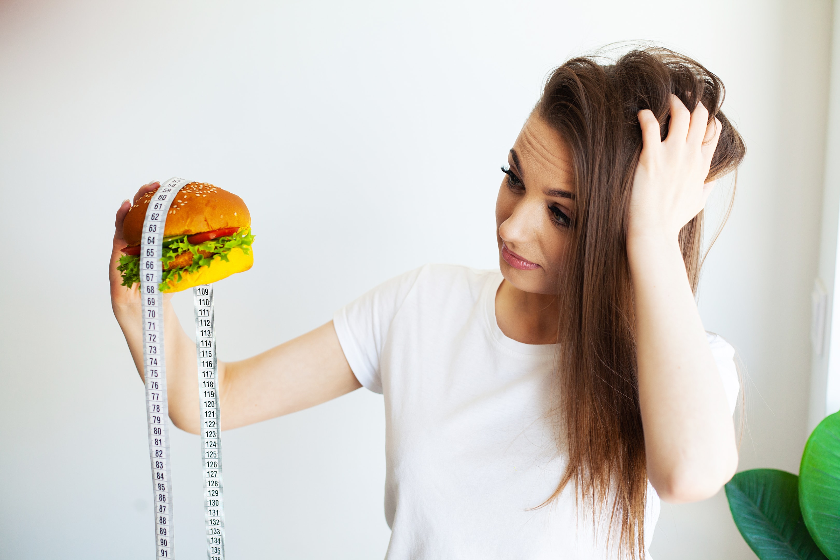 A woman looks stressed and confused while holding a burger wrapped in a measuring tape, illustrating the body image pressures and disordered thinking that can contribute to eating disorders in men.