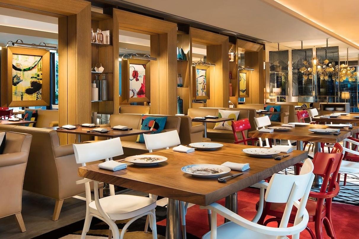  This contemporary dining room features thick wooden tables paired with white and red chairs, set against a backdrop of tan leather booths and light wood shelving. The space is decorated with vibrant abstract artwork and colourful throw pillows, creating a modern and welcoming atmosphere.