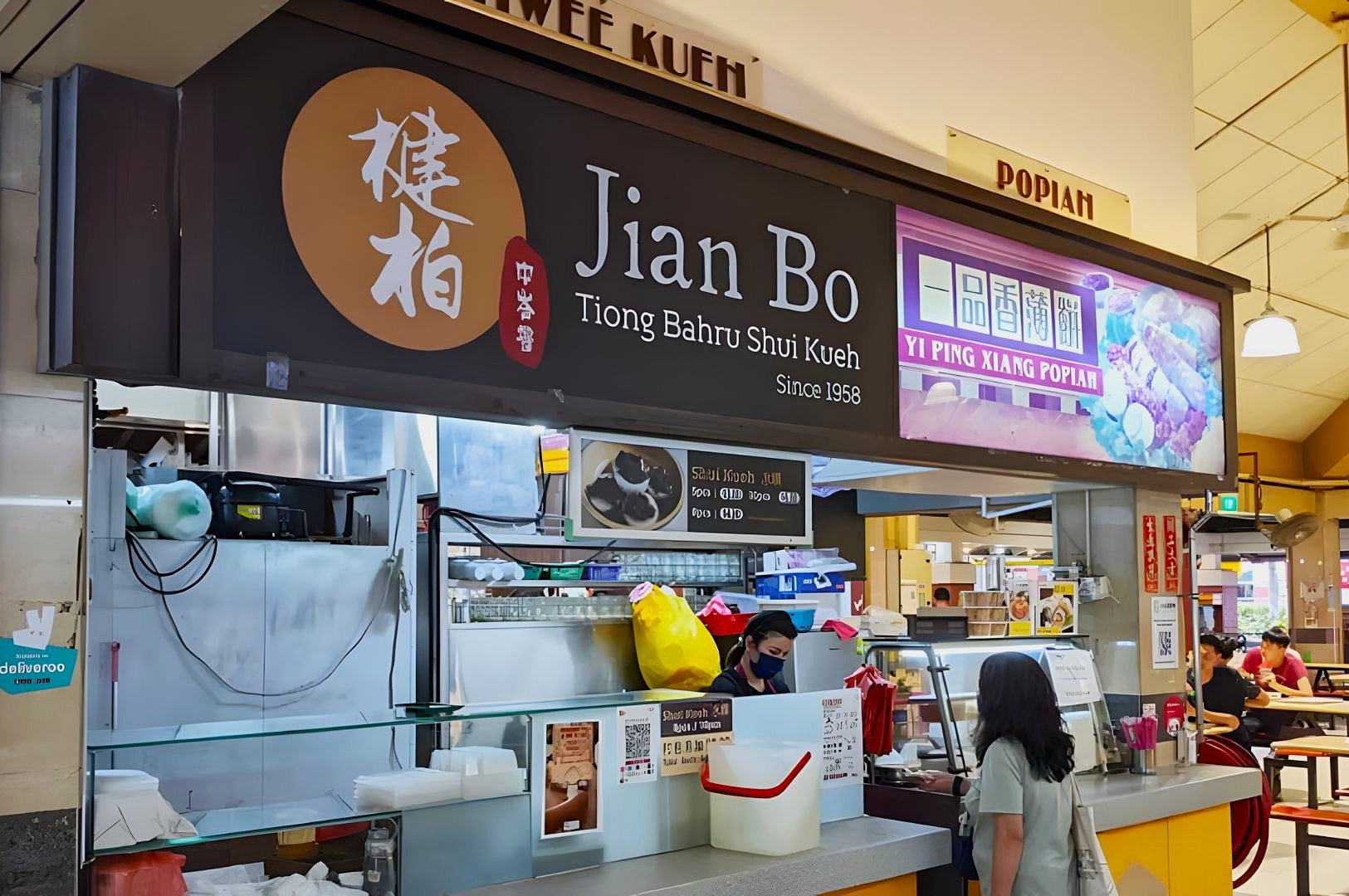 Stall front of Jian Bo Tiong Bahru Shui Kueh in a food court, featuring a large sign and colorful display. A person orders from a vendor.
