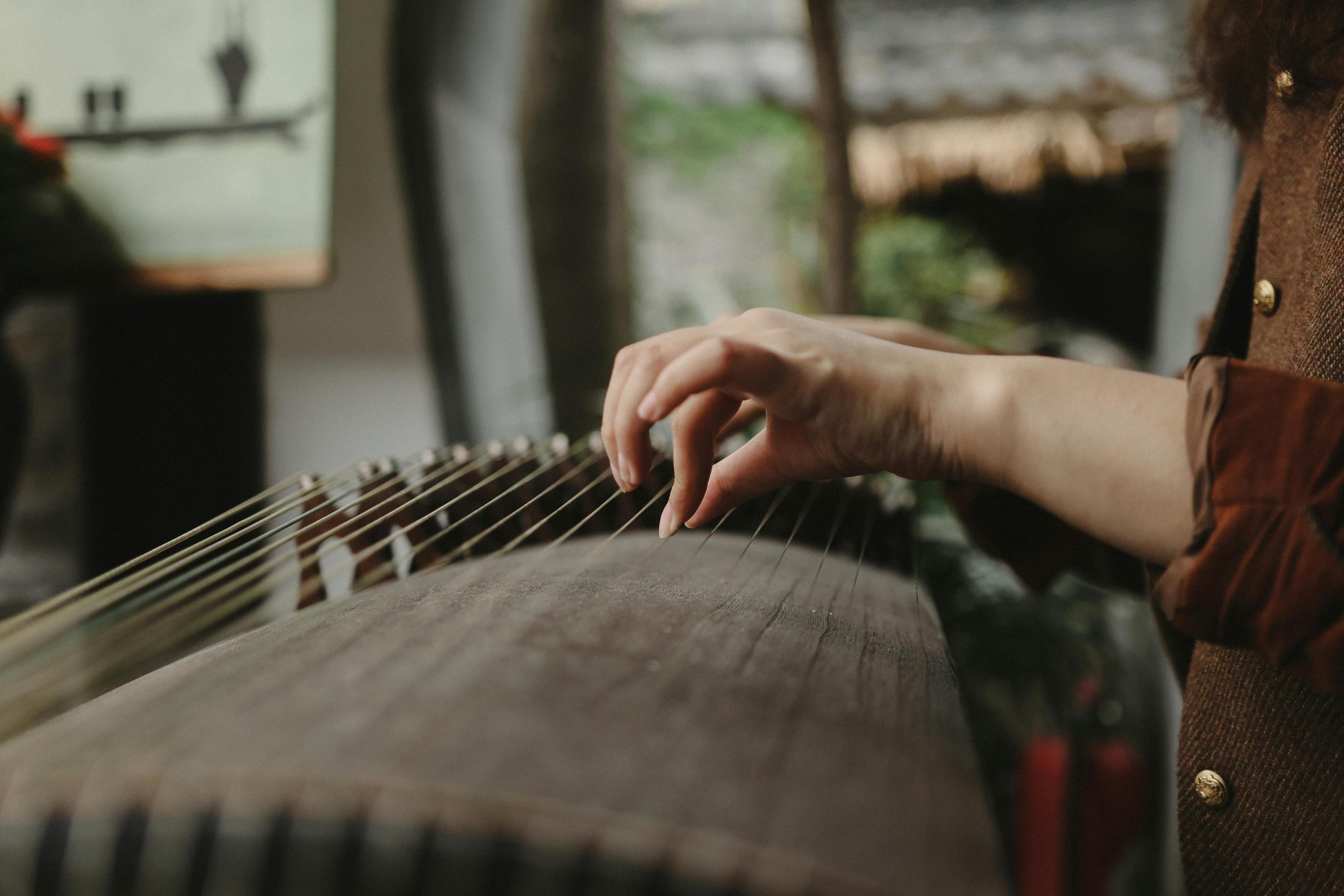 A close-up view focuses on a musician's hands delicately plucking the strings of a traditional wooden zither instrument. The performer is dressed in a brown jacket with brass buttons, playing against a softly blurred background that suggests an outdoor or cultural setting.