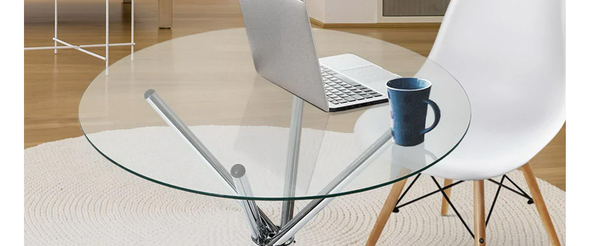 A glass coffee table, being used as a home office desk, with a laptop and a blue mug. Next to it is a white Eames DAW style dining chair. 
