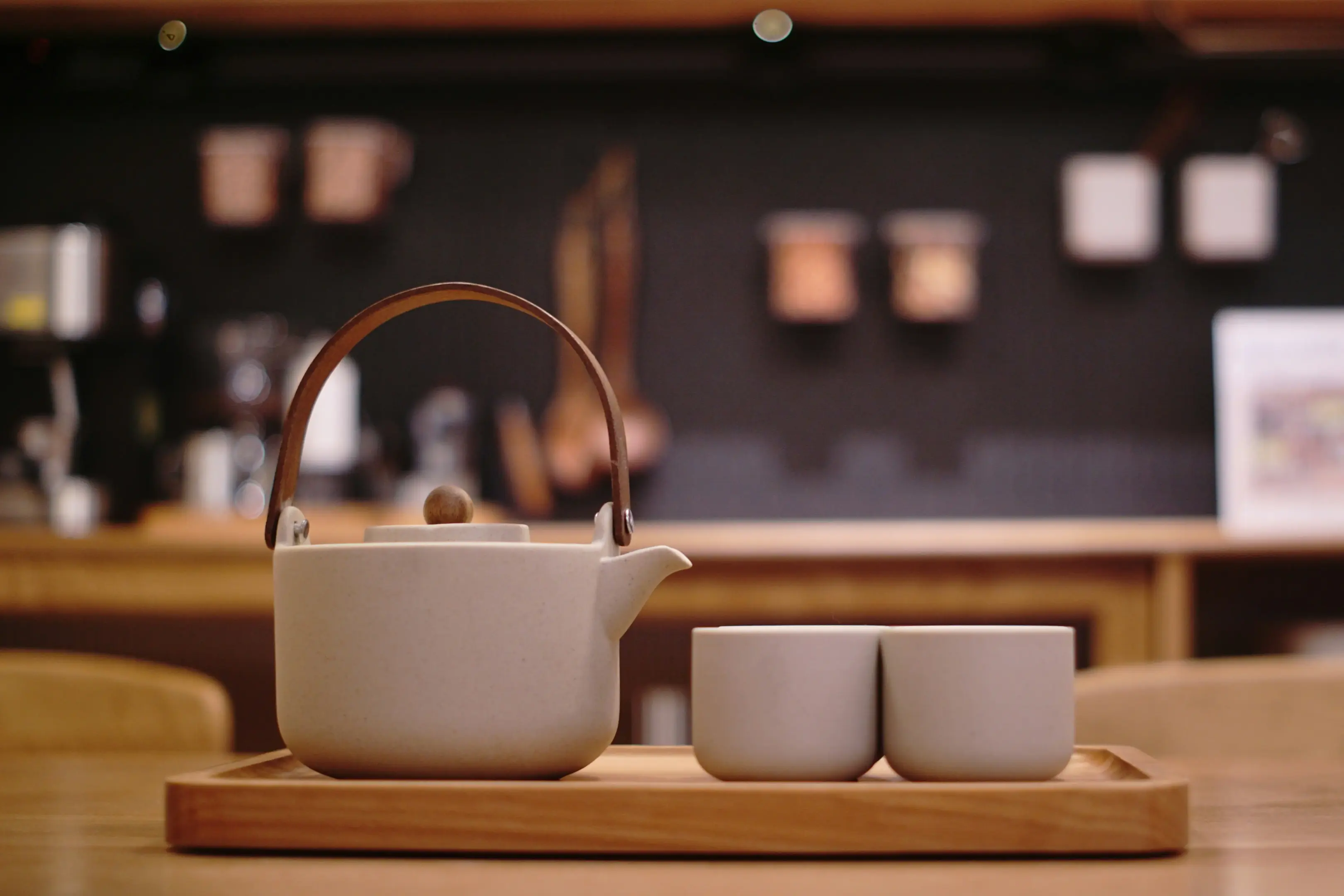 A minimalist ceramic tea set, featuring a light grey teapot with a bent wood handle and two matching cups, sits neatly on a wooden tray. The scene is captured with a shallow depth of field, softly blurring a warm kitchen background of wooden shelves and copper accents.