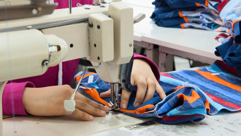 A sewing machine stitching striped fabric, with finished garments visible in the background.