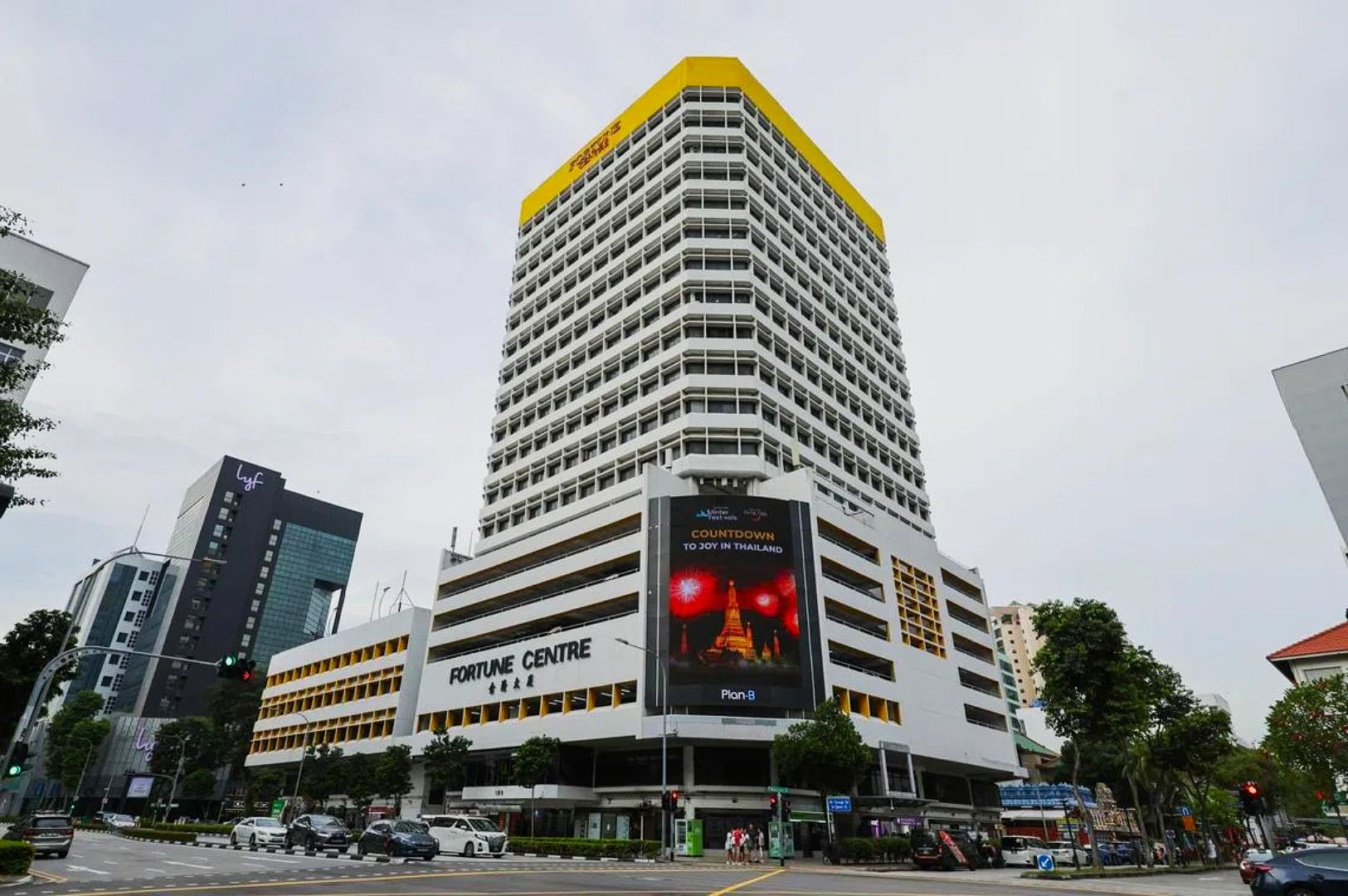 Multistory white building labeled "Fortune Centre" with yellow accents, large digital billboard displaying an ad featuring fireworks and countdown, street intersection in foreground.