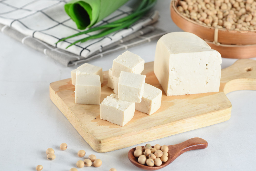 Cubes of tofu on a wooden board and a wooden bowl and spoon of soybeans. 