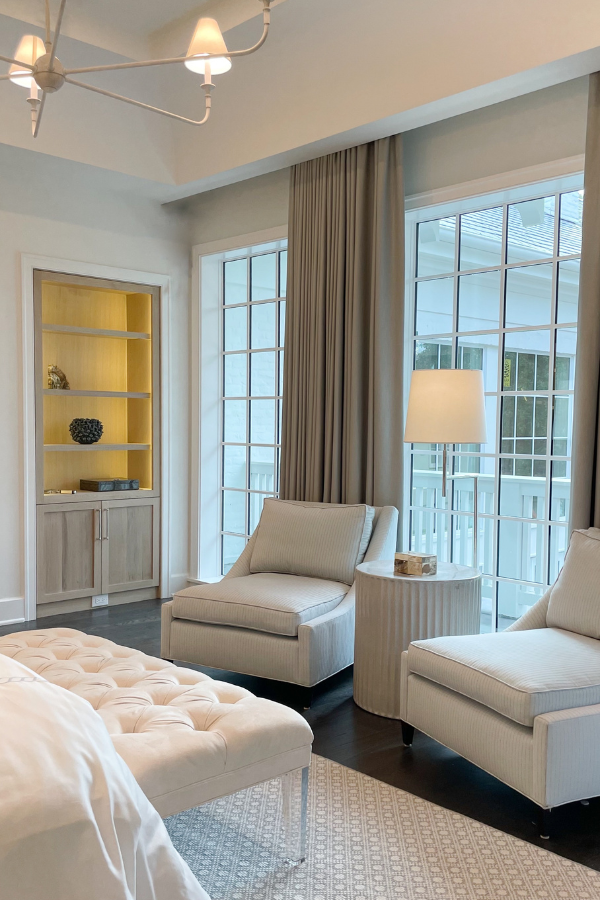 A serene sitting area with two upholstered lounge chairs, tall gray drapery, and a softly lit side table beside floor-to-ceiling windows.