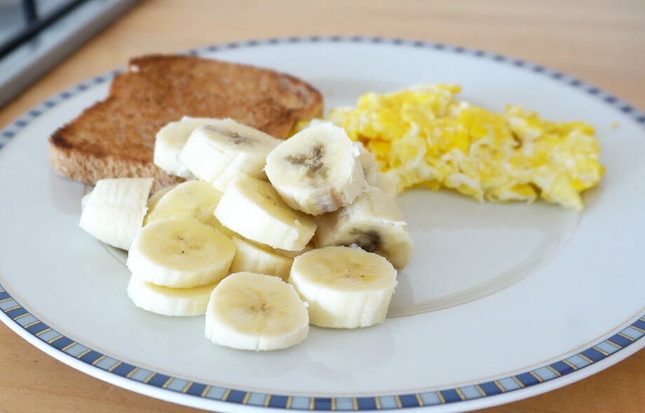 Piatto piano con sopra banana tagliata a fettine, uova strapazzate e pane tostato.