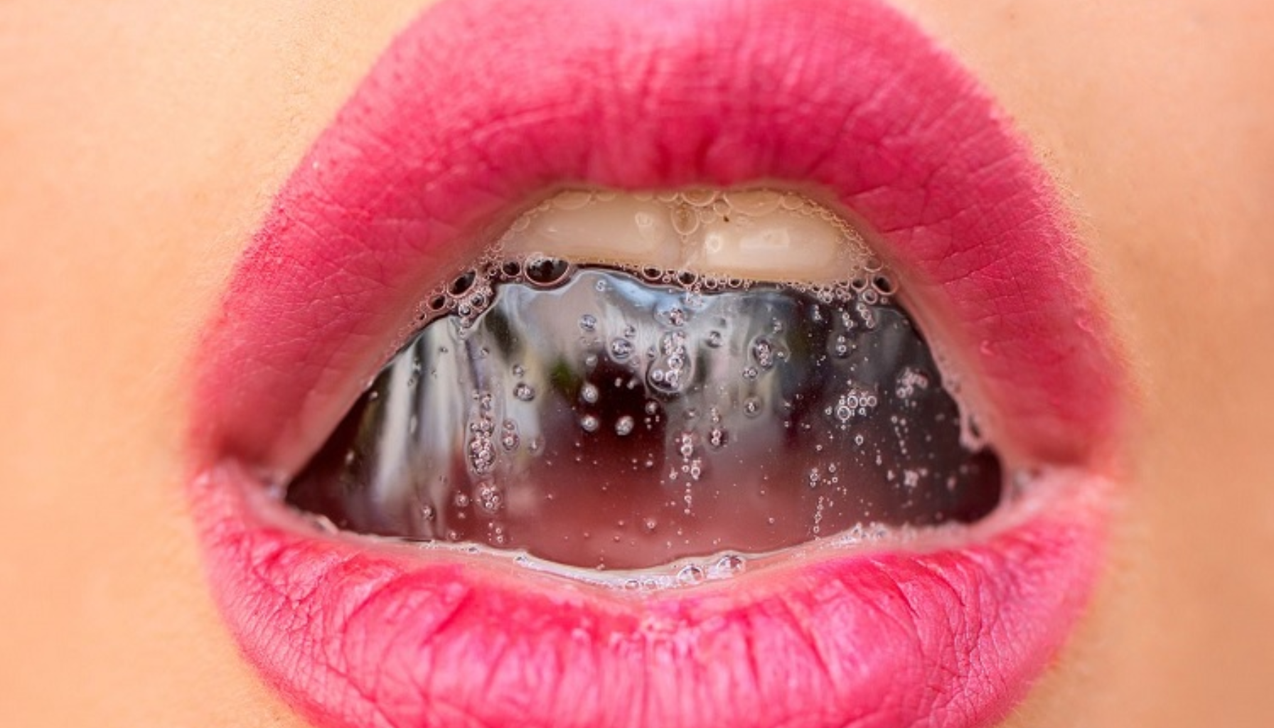 Nærbilde av lepper med rosa leppestift, viser speilrefleksjon med vanndråper og bobler i munnen.