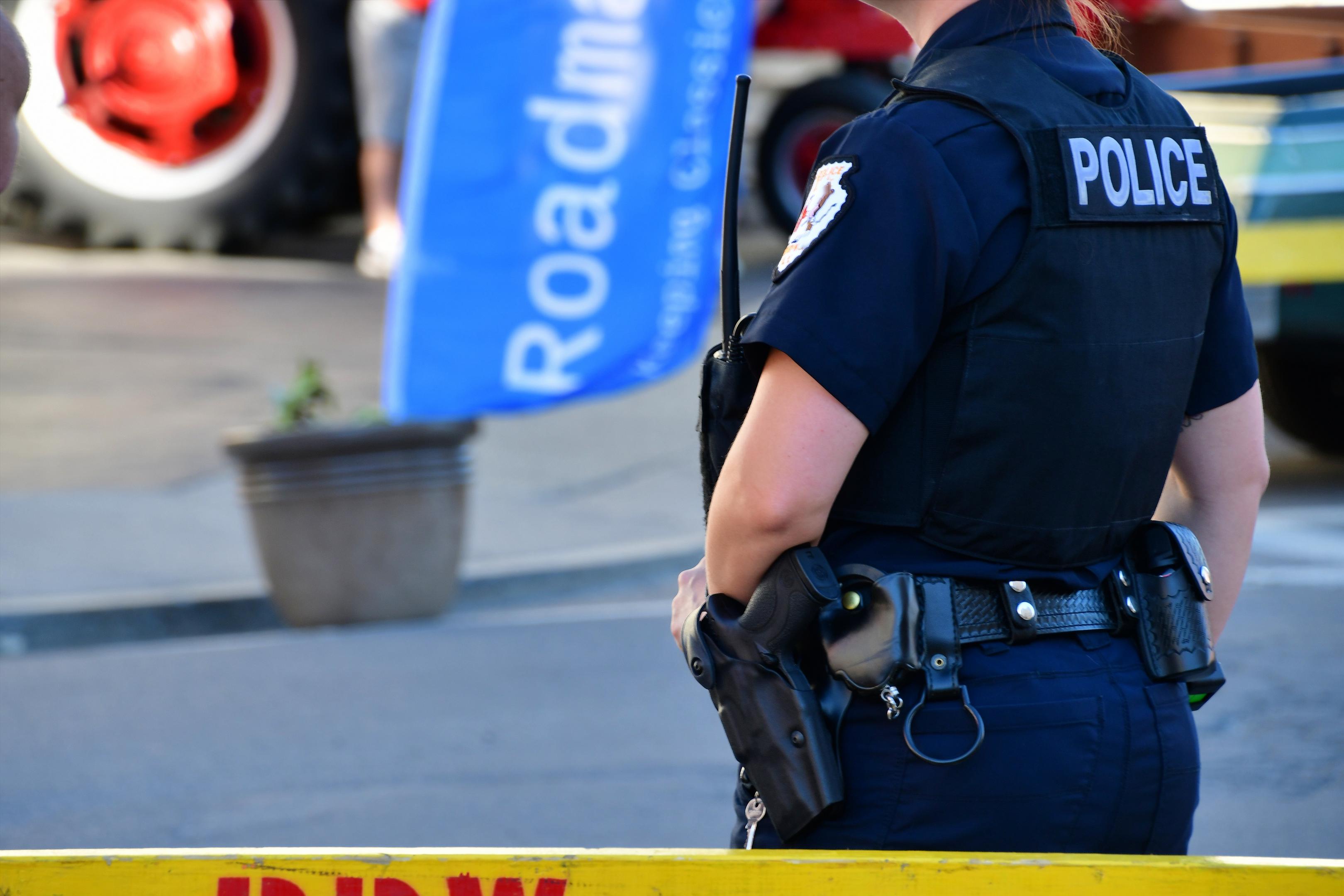 An officer stands in a dark blue uniform with black vest, a utility belt, and short sleeves. Visible on the back of the vest is the word "POLICE".