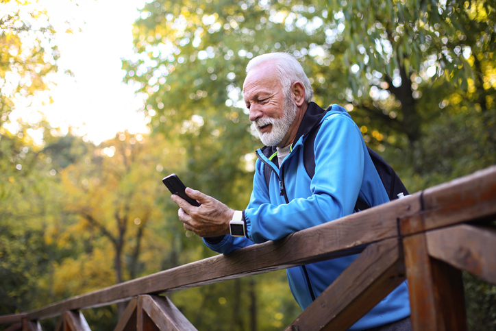 Older man with gray hair and a backpack checking his texts on a footbridge. 
