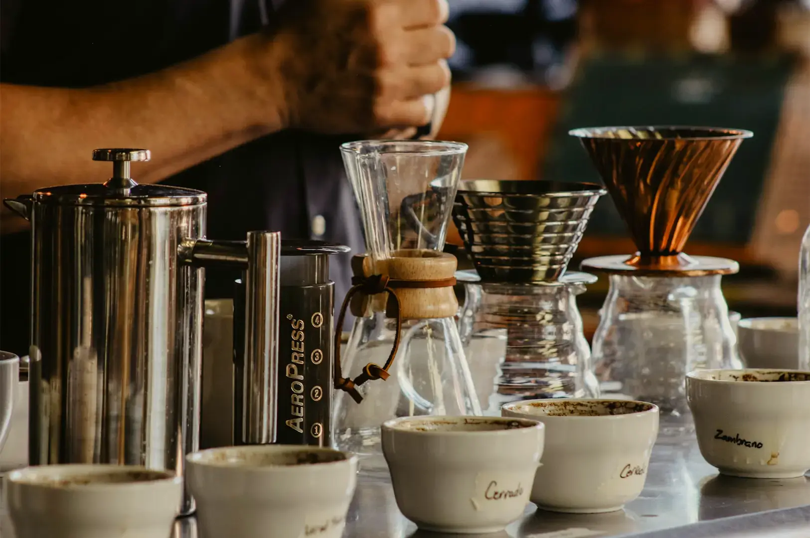 Coffee brewing items arranged in a line with white coffee cups below and a person preparing coffee behind.