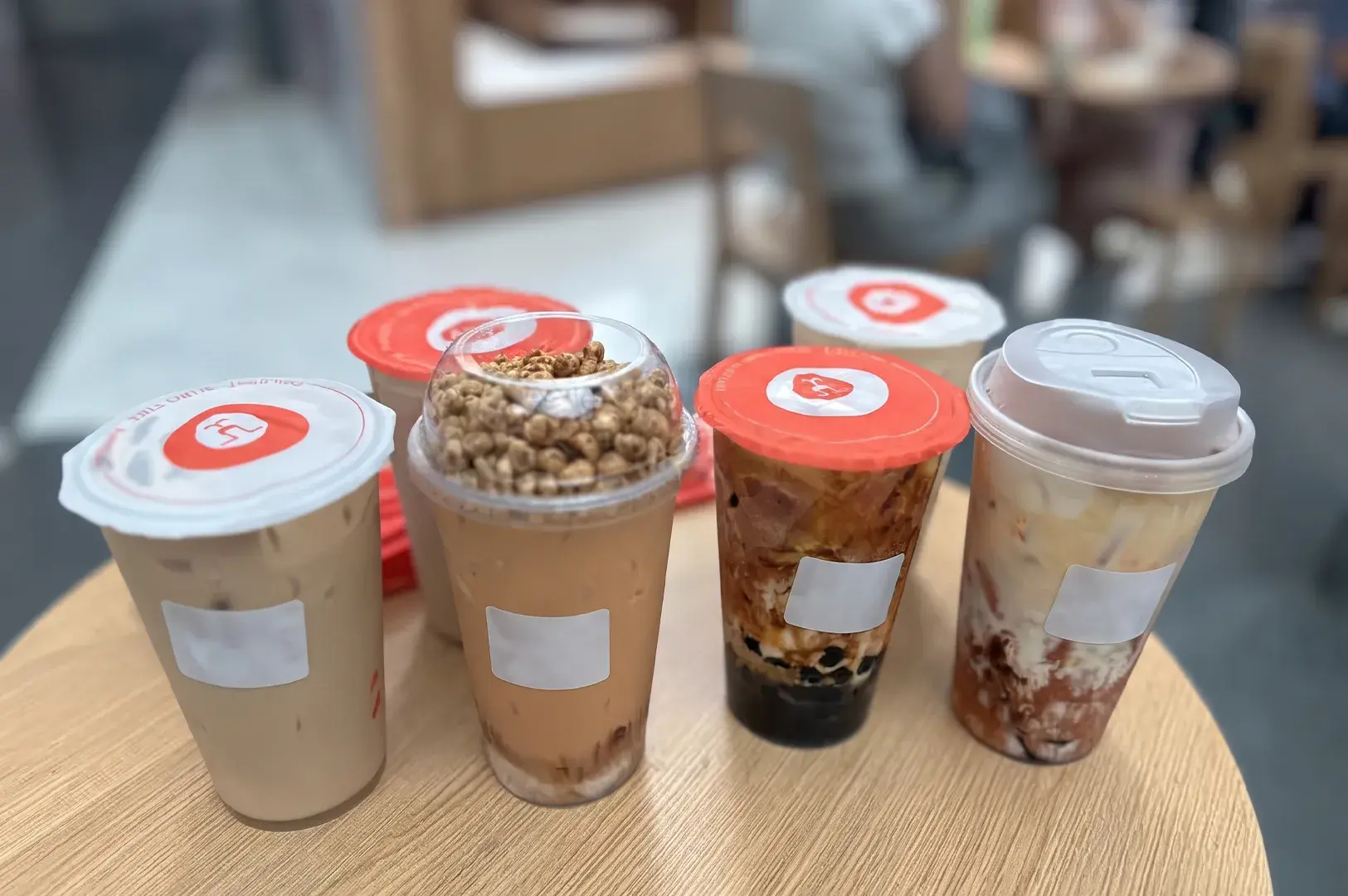 Six assorted specialty bubble teas and milk tea drinks arranged on a light round wooden table in a cafe.