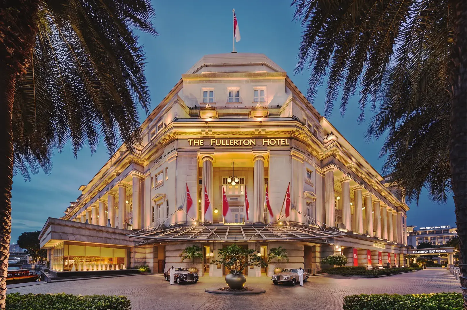 Grand Fullerton Hotel Singapore neoclassical architecture illuminated at night with palm trees and vintage cars.