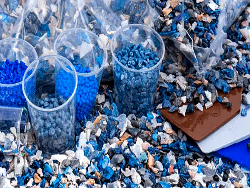 Small degradable plastic pellets in clear cups and colorful shredded plastic pieces scattered around them, in shades of blue, white, and gray