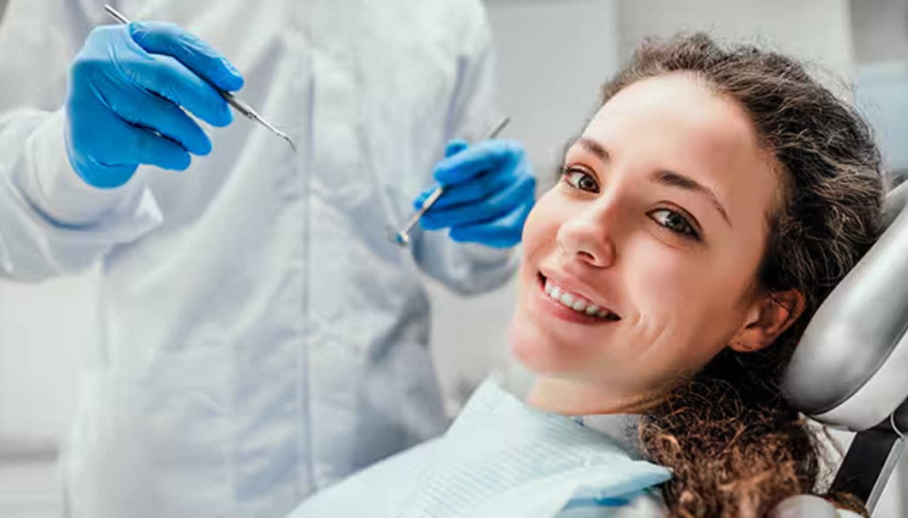 Trekke tann pris – Dette må du vite før behandling 15 Kvinne smiler mens hun sitter i tannlegestolen. Tannlegen i bakgrunnen holder tannverktøy og bruker blå hansker. Klar for undersøkelse.