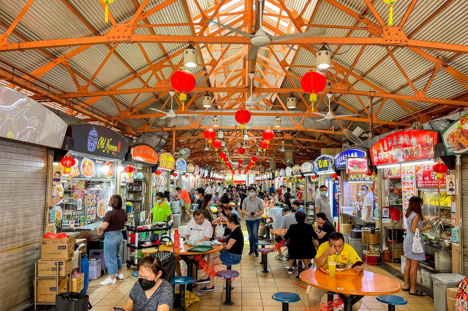 Interior view of a busy hawker center (like Maxwell or Lau Pa Sat) in Singapore, with people seated at tables under an orange roof and Chinese lanterns.