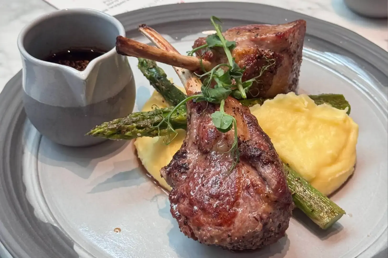 A grey ceramic plate presents two seared lamb chops resting over a bed of creamy mashed potatoes and grilled asparagus spears, garnished with fresh microgreens and accompanied by a small grey pitcher of dark jus.