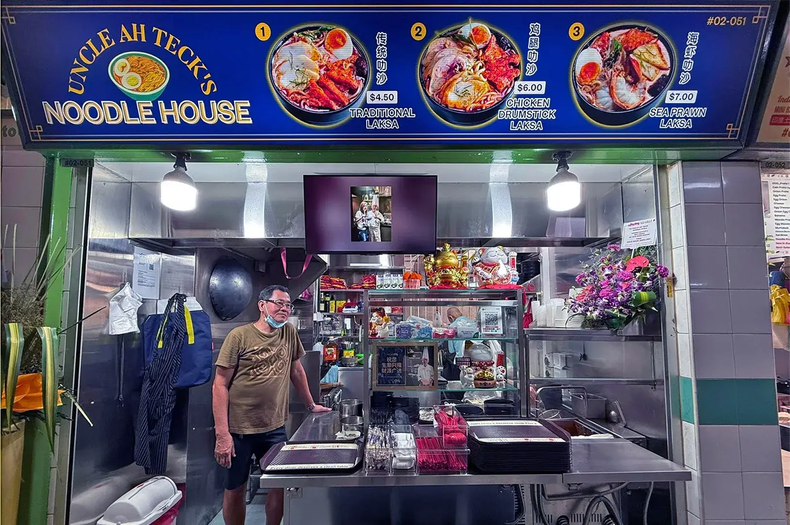 A noodle food stall named "Uncle Ah Teck's Noodle House." The interior has colorful menu signs displaying dishes like traditional laksa, with a man standing behind the counter, smiling warmly.
