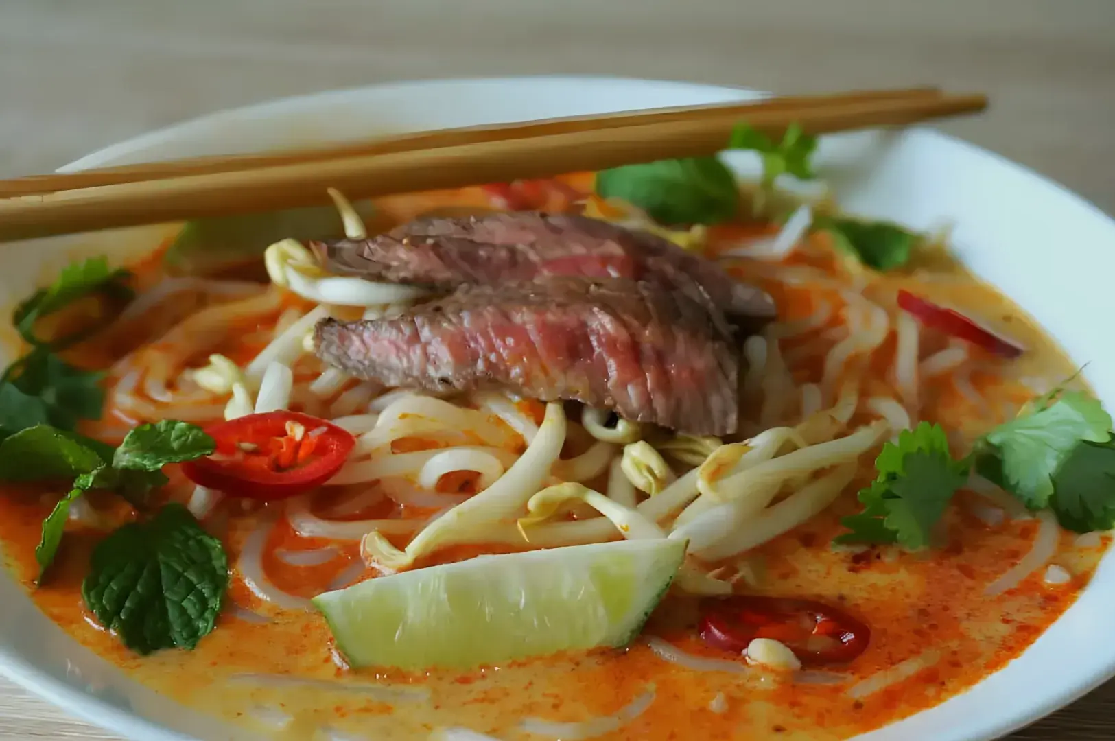A bowl of noodle soup topped with slices of beef, bean sprouts, fresh herbs, red chili slices, and a wedge of lime, with chopsticks resting on the edge.