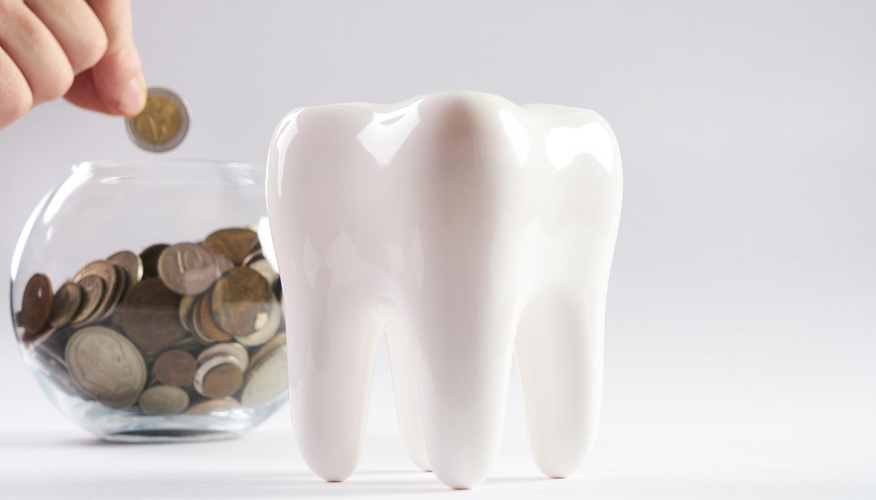 Tannhelseforsikring: En trygghet for økonomien 14 Hånd som legger mynt i glasskrukke ved siden av stor hvit tannformet sparebøsse på bord.